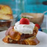 Two slices of challah french toast on a plate topped with tres leches syruc, whipped cream and a strawberry