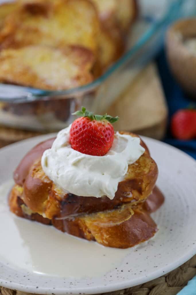 Two slices of challah french toast on a plate topped with tres leches syruc, whipped cream and a strawberry