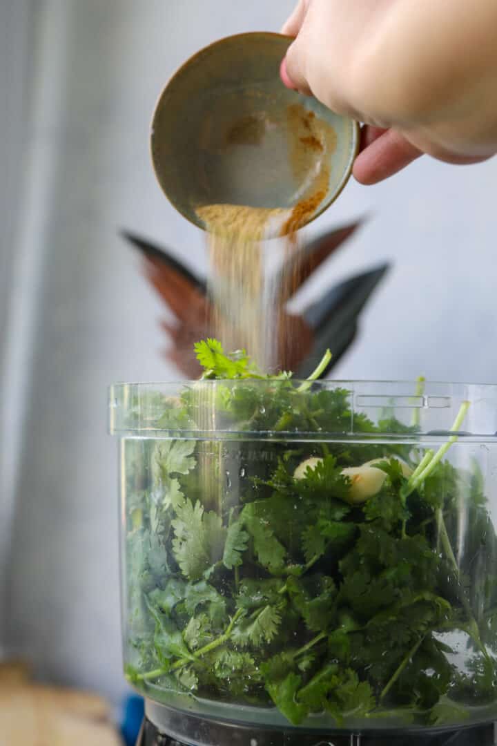 adding spices to a food processor filled with cilantro