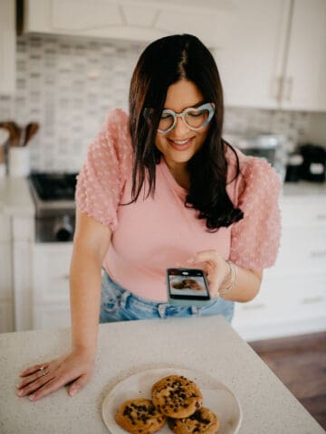 Cari Garcia taking a flat lay photo of a plate of chocolate chip cookies on a kitchen countertop with her phone.
