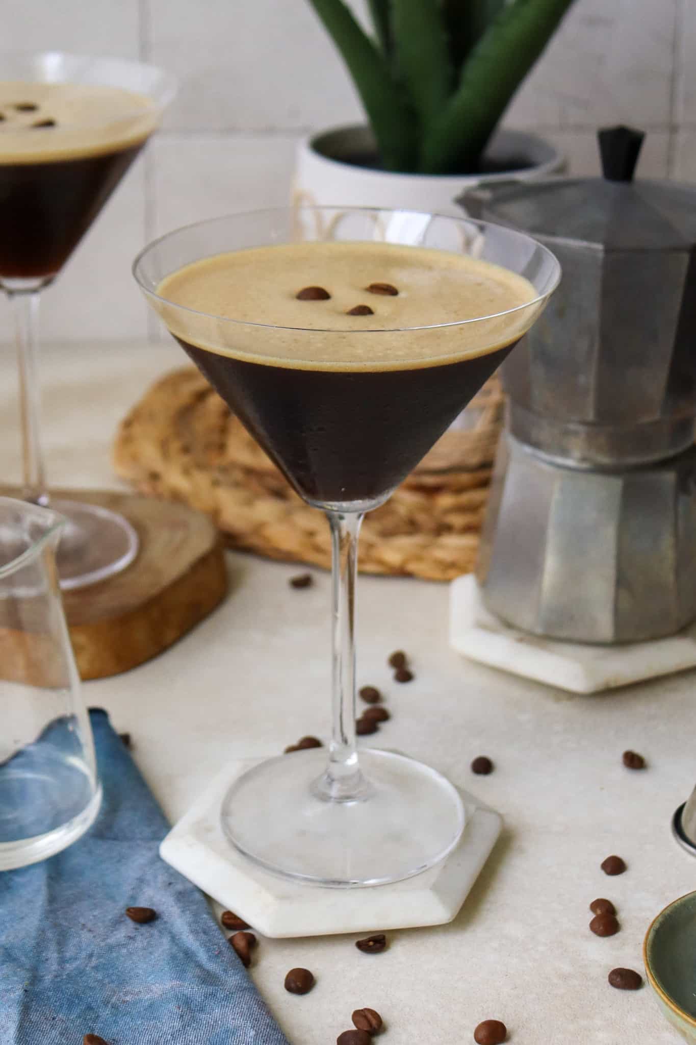 Cuban Espresso Martini in a martini glass placed on a hexagonal shaped coaster, placed on a white surface surrounded by coffee beans, an Italian espresso maker, another glass of the same martini and a blue tea towel.